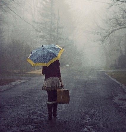 Mulher caminha por uma estrada, muito nublada, carregando uma mala na mão direita , ela está segurando um guarda-chuva azul e amarela, aberto.
