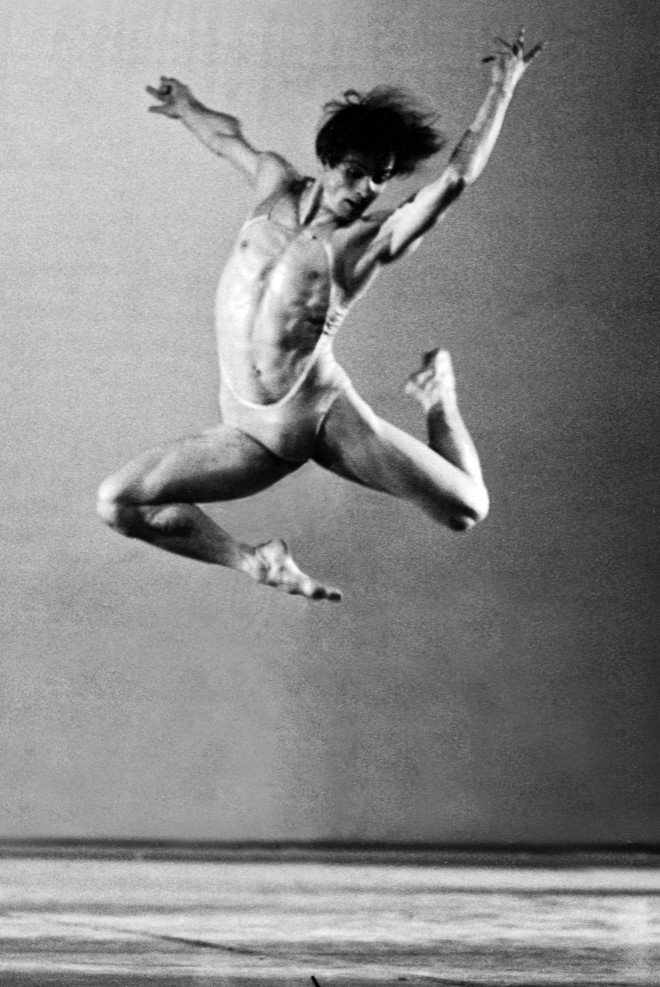 Rudolf Nureyev, bailarino, no palco, faz um salto, com a perna direita dobrada, perna esquerda , estendida para tras e dobrada , braço esquerdo estendido para cima, sobre a cabeça, braço direito estendido perpendicular ao corpo na altura do ombro e sua cabeça voltada para baixo. Ele usa um traje de ballet mínimo que apenas esconde suas partes íntimas.Seu salto, percebe-se está a mais de um metro do chão.