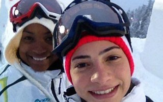 Lais de Souza, atleta brasileira, em imagem do rosto, está de sorrindo, com uma proteção na cabeça e um óculos para neve, ao fundo uma outra esquiadora também sorri. 