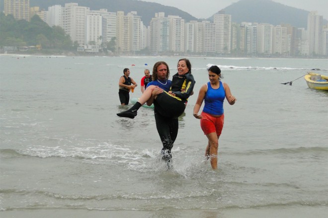 Mara Gabrilli , usando roupas esporte para surf, ela está nos braços de um outro atleta, também vestido com as roupas de mergulho e ao lado um outra jovem caminha, usando com bermuda vermelha e camiseta regata azul.