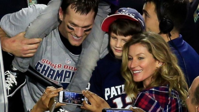 a foto mostra tom brady, gisele bundchem e o filho do casal no meio dos dois. ele está com uma criança nos ombros. todos felizes e sorrindo para foto na final do Super Bowl.