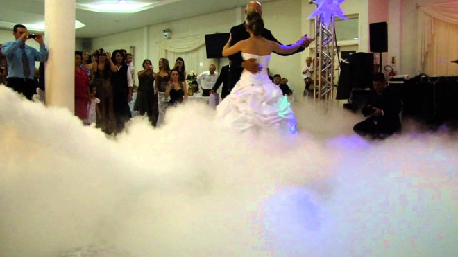 Na imagem uma noiva dança com o seu pai numa pista de dança da festa de casamento. Porém uma nuvem de gelo seco cobre a pista e cobre a noiva. Ao fundo os convidados fotografam esse momento.