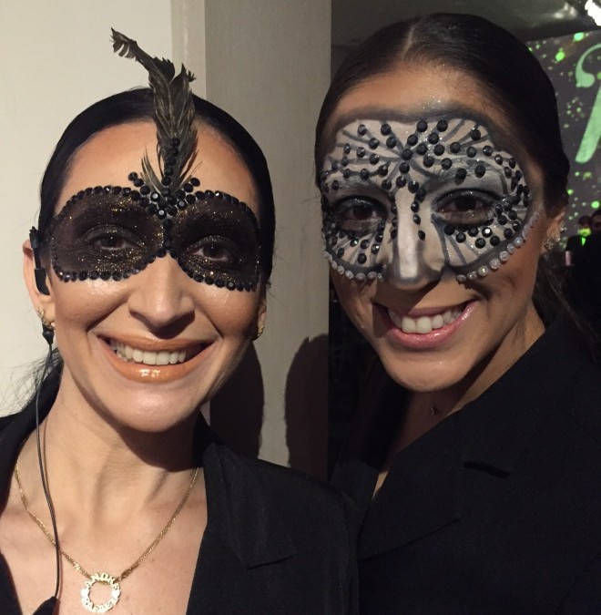 Duas moças de cabelos negros presos em um coque vestem preto e estão sorrindo para a câmera. As duas estão com uma máscara de maquiagem negra pintada sobre a pele. Nas bordas da pintura pedras e cristais brilhantes enfeitam a máscara e o efeito é muito bonito e real.
