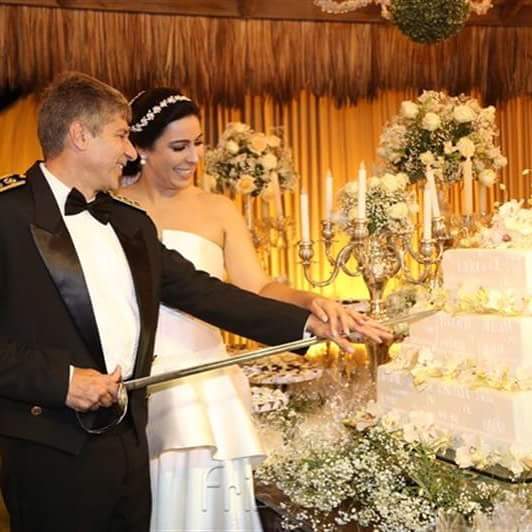 Os noivos, Lacira e Gil, ele de smoking ela de vestido branco tomara que caia estão prestes a cortar o grande bolo quadrado de casamento de 4 andares. Ele empunha a espada para cortar como é tradição em casamentos de militares.