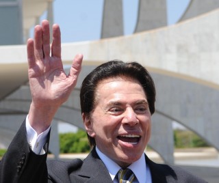 O Apresentador Silvio Santos, ele está sorrindo, com cabelos escuros. Ele veste um terno escuro , camisa branca, gravata escura com detalhes em tons escuros.